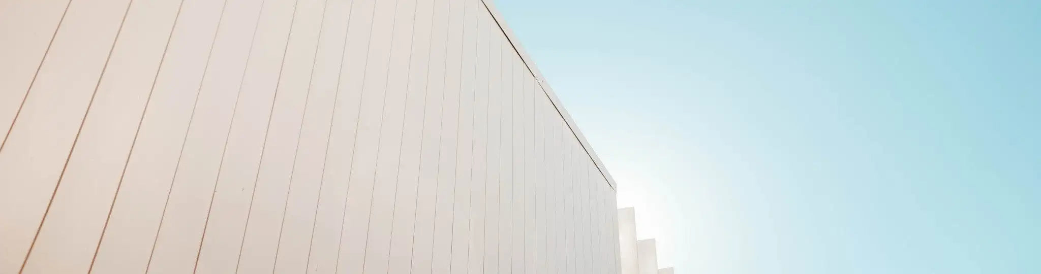 The image shows a modern, minimalist building facade with vertical white siding panels under a clear blue sky.